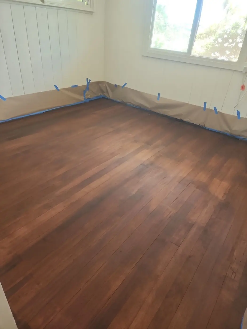 Hardwood floor refinish: wider room view showing the finished planks corner to corner, kraft-paper masking still in place along the walls