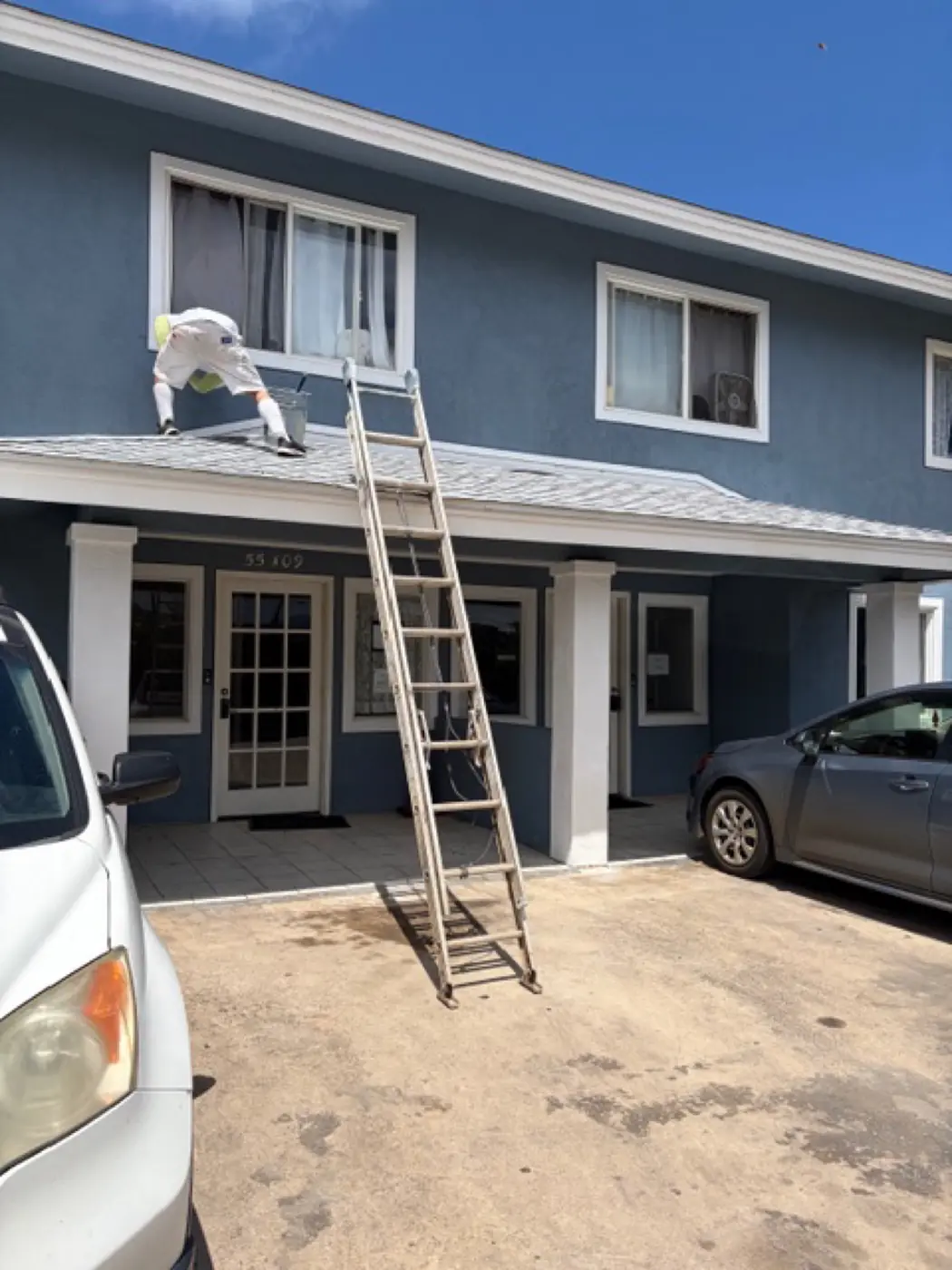 Salt-air spec mid-project: ladder propped against the two-story exterior, salt-spec primer going on the slate-blue base coat, paint truck staged at the curb